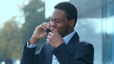 Resmi giyinmiş Afro-Amerikalı bir adam dışarıda duruyor, gülümsüyor ve modern bir şehir arka planına karşı bir telefon görüşmesi yapıyor..