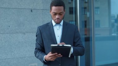 Resmi giyinmiş Afrikalı Amerikalı bir adam, çağdaş bir ofis yapısının yanında duran bir cihazla ilgili bilgileri gözden geçiriyor..