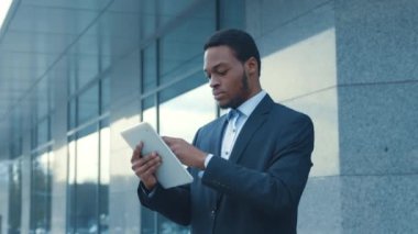 Takım elbiseli Afro-Amerikalı bir adam öğleden sonra bir cam ofis binasının önünde dururken tabletle etkileşime giriyor..