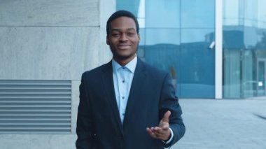 Profesyonel bir Afro-Amerikalı takım elbiseli adam güneşli bir günde elini modern bir ofis binasının önüne uzatıp gülümser ve jest yapar..