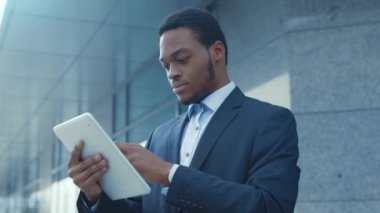 Takım elbiseli Afrikalı Amerikalı bir adam, çağdaş bir ofis binasının önünde dururken tabletine odaklanır..