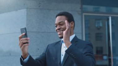 Takım elbiseli Afro-Amerikalı bir genç, şehir dışında bir video görüşmesi yaparken gülümsüyor..