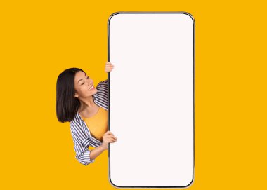 Harika bir teklif. Heyecanlı Asyalı bir kadının portresi büyük, dikey bir cep telefonundan beyaz beyaz ekranlı ve cihaz ekranına bakıyor. Boş boş alanı olan Gadget modeli, sarı turuncu duvar