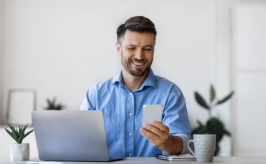 Yakışıklı erkek ofis yöneticisi iş yerinde akıllı telefon kullanıyor, masada oturuyor, sosyal medyayı kontrol ediyor veya uygulamayı tarıyor, mesaja cevap veriyor, ara veriyor, ekrana bakıyor ve gülümsüyor, boşluğu kopyalıyor.
