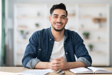 Keyifli Arap adam çalışma masasında oturuyor ve kameraya gülümsüyor, ofis içi, fotokopi odası. Orta Doğulu mutlu genç adam elinde defter ve kağıtlarla masada oturuyor.