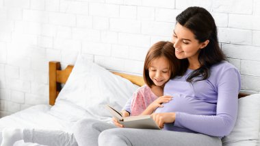 Loving pregnant mom reading book to her little daughter while resting together in bed, copy space