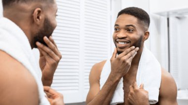 Çıplak gövde dokunaklı sakalı ile mutlu afro adam banyoda aynaya bakıyor. Banyo konseptinde modern adam