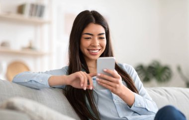 Hafta sonu eğlencesi. Gülümseyen Asyalı kız internette akıllı telefonuyla geziniyor, oturma odasında kanepede dinleniyor, boş yer var.