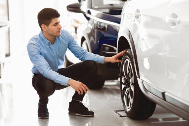 Muayene arabası. Yepyeni bir arabanın yanında oturan yakışıklı adam tekerlek jantlarını ve lastiklerini kontrol ediyor.