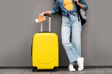 Unrecognizable woman in stylish casual outfit traveller standing over grey background, carrying yellow luggage and backpack, holding passport and flight tickets, cropped, copy space
