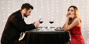 Failed Date. Unhappy young woman sitting alone while her boyfriend stuck into his phone during dinner in restaurant, side view with free space