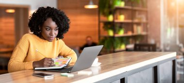 Concentrated black girl browsing job opportunities on laptop at cafe, writing in notepad