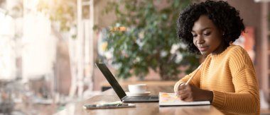 İnternette yeni bir iş arayan motive olmuş afro genç kadın, kafede dizüstü bilgisayar kullanıyor, notlar alıyor, yan görüş alıyor, boşlukları kopyalıyor.