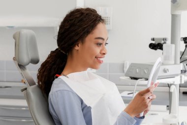 Modern klinikte stomatolojik tedaviden sonra beyaz gülüşünü kontrol eden güzel siyah kadın.