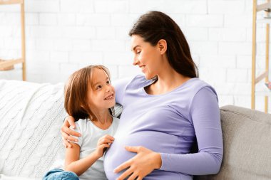 Neşeli hamile kadın küçük meraklı kızıyla konuşuyor, oturma odasında koltukta oturuyor, boşluğu kopyalıyor.