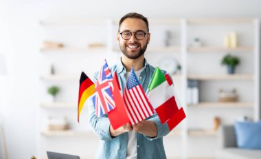 Modern education and student exchange. Cheerful young Caucasian guy holding bunch of different flags at home. Happy millennial man advertising foreign languages school, emigrating overseas clipart