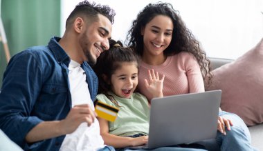 Bilgisayarı ve kredi kartı olan neşeli Arap ailesi evdeki koltukta dinlenirken internetten alışveriş yapıyor mutlu bir orta doğulu ebeveyn ve internetten alışveriş yapan küçük bir kız çocuğu.
