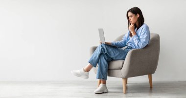 Dizüstü bilgisayar ekranına bakan, koltuğunda oturan, online çalışmayla sorunu olan ya da beyaz stüdyo duvarına karşı çalışan, afişi kopya alanı olan ciddi genç bir kadın.