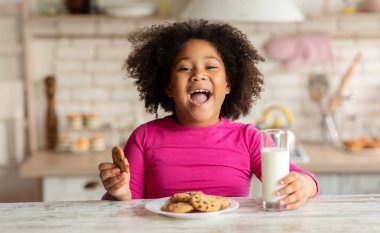 Neşeli küçük siyah kız, masada oturmuş süt ve kurabiyelerin tadını çıkarıyor, tatlı Afro-Amerikan çocuğu mutfakta atıştırıyor, sağlıklı kalsiyum içeceği ve ev yapımı pastayla bir ısırık alıyor,