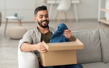 Hızlı teslimat hizmeti konsepti. Mutlu genç bir Arap paket alıyor, karton kutuları açıyor, oturma odasındaki kanepede oturuyor. Orta Doğulu erkek alıcı çevrimiçi alışveriş alışverişinden memnun