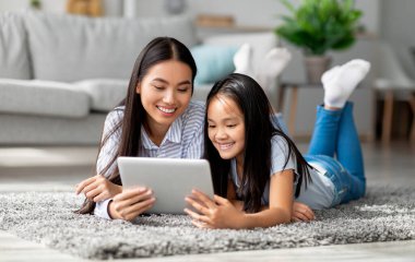 Heyecanlı Asyalı anne kızına dijital tablet dersinde yardım ediyor oturma odasındaki halının üzerinde birlikte yatıyor. Genç anne ve çocuğu birlikte online ev ödevi yapıyorlar.