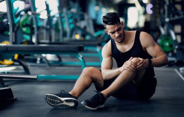 Spor Travması. Spor salonunda yaralı dizine dokunan genç Arap erkek sporcu, spor salonunda antrenman yaptıktan sonra yerde oturan ve bacağı yaralanan orta doğulu adamı üzüyor.