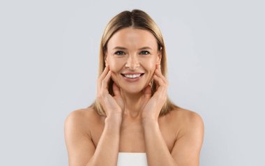 Face injections concept. Pretty blonde middle aged woman wrapped in white bath towel touching her face skin and smiling, enjoying results of face care treatment, grey studio background