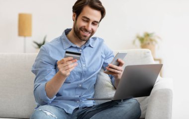 Memnuniyet duyulan adam cep telefonu ve kredi kartı kullanıyor dizüstü bilgisayarla oturuyor ve evde alışveriş yapıyor. Akıllı Telefonlu Müşteri Dükkanları. Mobil Bankacılık Uygulaması ve Ödeme Doğrulama Konsepti