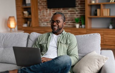 Happy adult african american male in glasses watching video on laptop, study, enjoy rest at free time alone in living room interior. Social media chat remote, new normal with technology, work at home