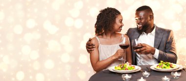 Young afro couple in beautiful outfit having festive dinner at restaurant, drinking red wine, eating healthy food