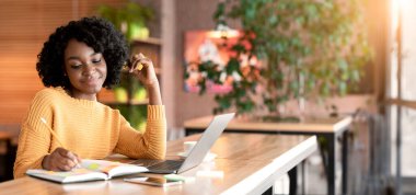 Dreamy black girl writing down thoughts while spending time at cafe, using laptop, copy space