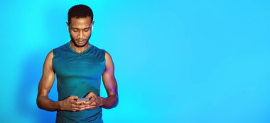 Sporty Black Man Telefon ve Kulaklık Kullanıyor Mavi Stüdyo Arkaplanda Duran Kulaklıklar. Fitness için çalma listesi. Boşluğu Kopyala