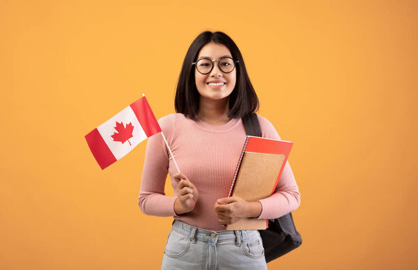 Travel, modern education and student exchange. Young cheerful asian woman in glasses with notebooks, backpack holding small flag of Canada, isolated on beige background, studio shot, empty space