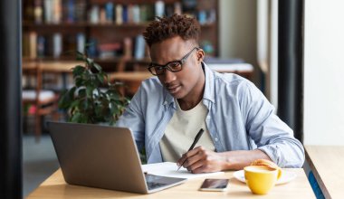 Ciddi siyahi adam şehir kafesinde dizüstü bilgisayarın önünde ders çalışırken not alıyor.