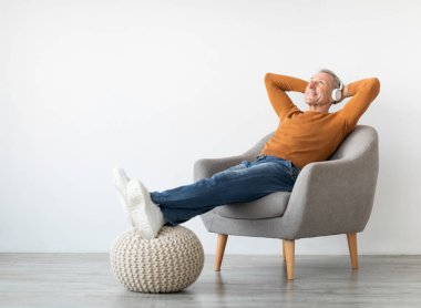 Dinlen ve rahatla kavramı. Sakin, olgun bir adam arkasını yaslanmış, müzik dinliyor, sesli kitap ya da podcast dinliyor, kablosuz kulaklıklarla ve beyaz duvarla meditasyonun keyfini çıkarıyor.