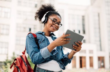 E-öğrenme. Disarida Modern Üniversite Binasi 'nin yakinlarinda duran kulaklik takan Tablet Bilgisayar kullanan mutlu siyah kadin ögrenci. Genç kız dışarıda internet gezintisi yapıyor. Modern Çevrimiçi Eğitim
