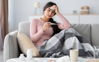 Ekoseli üzgün Hintli kadın, gripten muzdarip, kanepede oturuyor ve oturma odasındaki termometreye bakıyor. Soğuk algınlığı, ateş, evde hastalık tedavisi ve covid-19 tecrit, boş alan