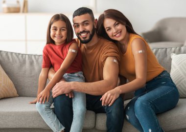 Coronavirus Vaccination Advert. Happy Vaccinated Family Of Three People Showing Arm With Sticking Plaster After Covid-19 Vaccine Vax Injection Posing Sitting On Couch In Living Room, Smiling To Camera clipart