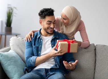 Elinde tesettürlü, mutlu Müslüman bir kadın. Şaşırmış sevgilisine sarılıyor. İç mekan, boş alan. Duygusal bir kadın, neşeli kocasını kucaklıyor, Sevgililer Günü için hediye kutusu veriyor.