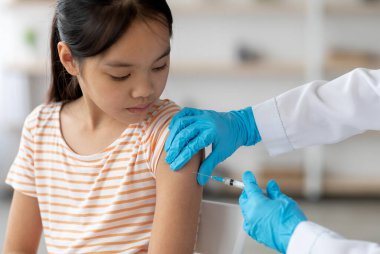 Unrecognizable doctor hands in protective gloves making vaccination against coronavirus of flu for little asian girl, home interior, closeup shot. Kids immunization against various disease concept