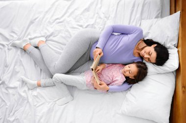 Genç bir kadının, yatakta dinlenirken küçük kızına kitap okumasını beklemesinin en iyi görüntüsü.