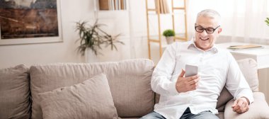 Halk ve Teknoloji Konsepti. Gözlüklü olgun bir adamın evdeki koltukta oturup akıllı telefon kullanmasına sevindim.