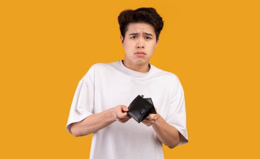 Meteliksizim. Asyalı üzgün iflasın portresi boş cüzdanını gösteriyor ve kameraya bakıyor. Ağlayan erkek model turuncu stüdyo arka plan duvarında tek başına duruyor. Yoksulluk Kavramı.