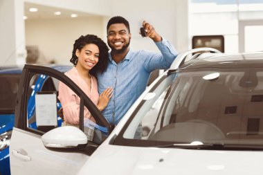 Cheerful Afro Spouses Standing Near New Car Showing Key Smiling To Camera In Auto Dealership Center.