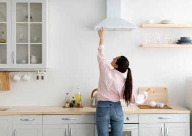 Ev kavramı. Arka planda, sıradan bir bayanın menzil egzoz kaputunda seçtiği ve seçtiği mod, ocağın üstündeki mekanik vantilatörün düğmesine basıyor. Evdeki modern mutfakta yemek pişiren kadın.