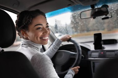 Harika bir yolculuk, araba yolculuğu ve şehirde iyi yolculuklar. Mutlu genç Afrikalı Amerikalı kadın sürücü arka koltukta yolcu koltuğuna dönüyor. Şehir yolu görüntüsü, yağmur damlalı pencere ve akıllı telefon, boşluğu kopyala