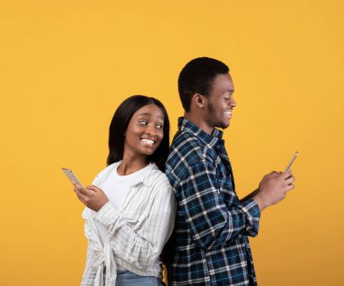 Bloglar, sosyal medya, sıradışı mesaj ve ilginç olay. Neşeli, Afro-Amerikalı genç bir bayan gülümseyen kocanın akıllı telefonuyla omzunun üzerinden bakıyor, sarı arka planda izole edilmiş, stüdyo çekimi.