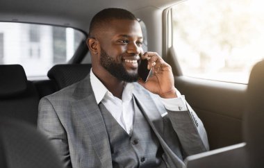 Bilgisayarlı mutlu siyah iş adamı müşterileriyle cep telefonuyla konuşuyor, pencereden bakıyor, iş toplantısına arabayla gidiyor, Afrikalı yönetici arka koltukta oturuyor, telefonla konuşuyor.