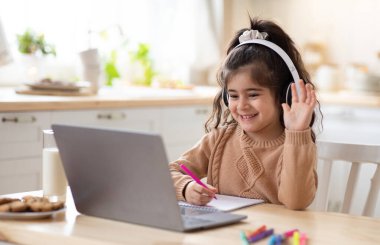 Mesafe öğrenme. Sevimli küçük ev okulu öğrencisi mutfakta dizüstü bilgisayarla video dersi alıyor, Coronavirus karantinası sırasında bilgisayarla çalışıyor, online eğitimin tadını çıkarıyor, kameraya el sallıyor.