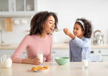 Kahvaltı zamanı geldi. Gülümseyen Afrikalı Amerikalı küçük bir kız genç bir kadını mısır gevreğiyle besliyor, annesiyle lezzetli bir kahvaltı yapıyor, süt içiyor, sabahları birlikte vakit geçiriyor.